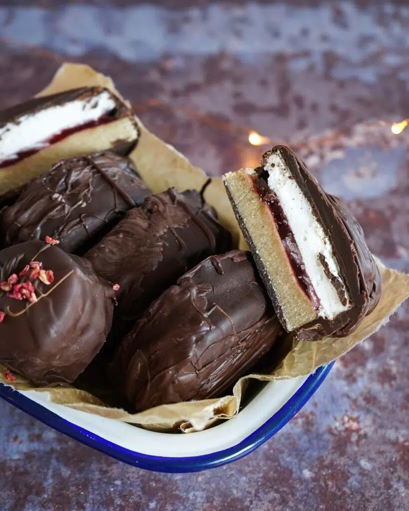 A dish of mini vegan wagon wheels cut in half to show the layers of vegan marshmallow, tangy raspberry jam and oat biscuit coated in dark chocolate