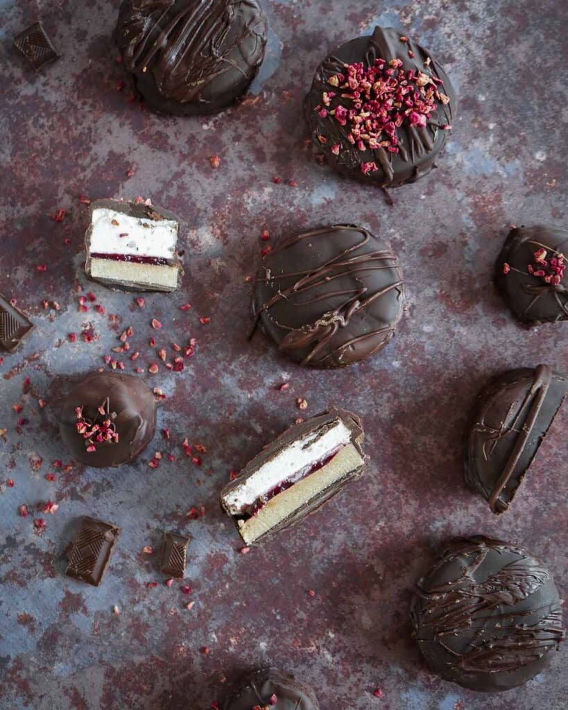 Mini vegan wagon wheels cut in half to show the layers of vegan marshmallow, tangy raspberry jam and oat biscuit coated in dark chocolate