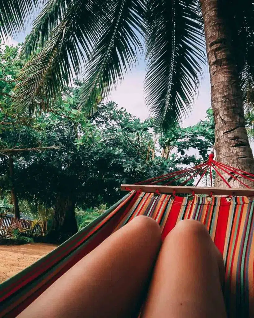 A woman sat on a striped red hammock underneath a palm tree on a beach in Vav'au Tonga