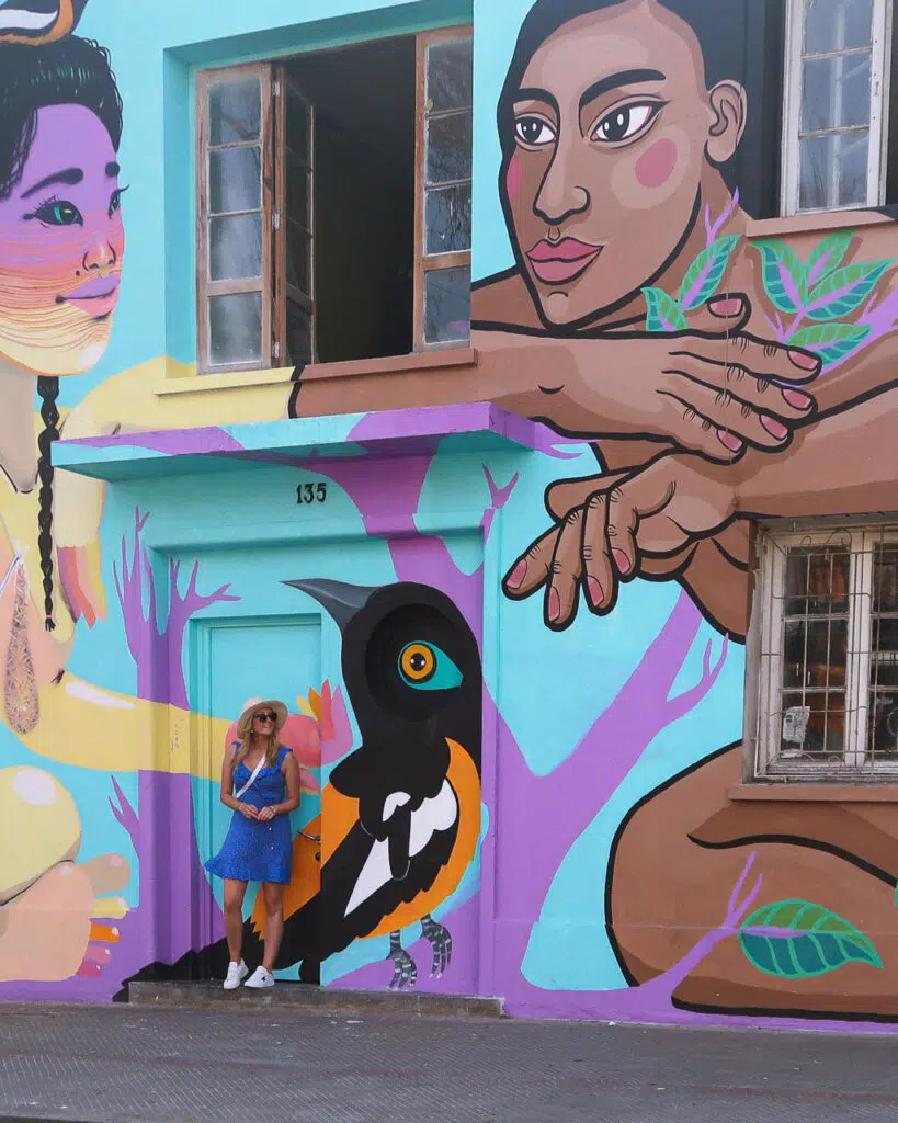 A woman in a blue dress stood outside a brightly coloured building, painted with street art depicting humans and birds