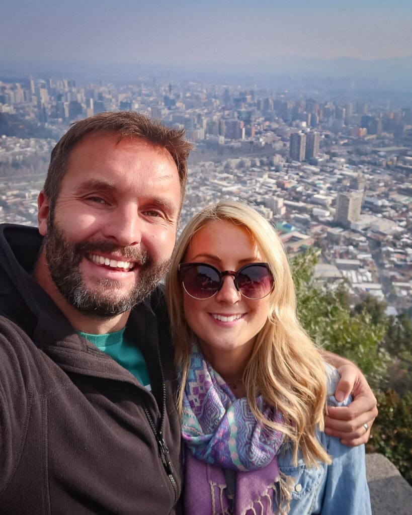 A happy couple smiling at the camera with the view of the city of Santiago, Chile behind them.