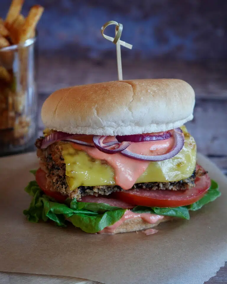 A stacked vegan fish burger with melty cheese, chilli mayo, red onion, lettuce and tomato in a soft bun
