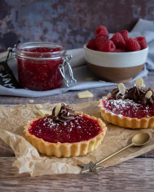 Two pretty vegan jam tarts in golden pastry cases, topped with piped chocolate cream and set on a worktop with a pot of raspberry jam and fresh raspberries