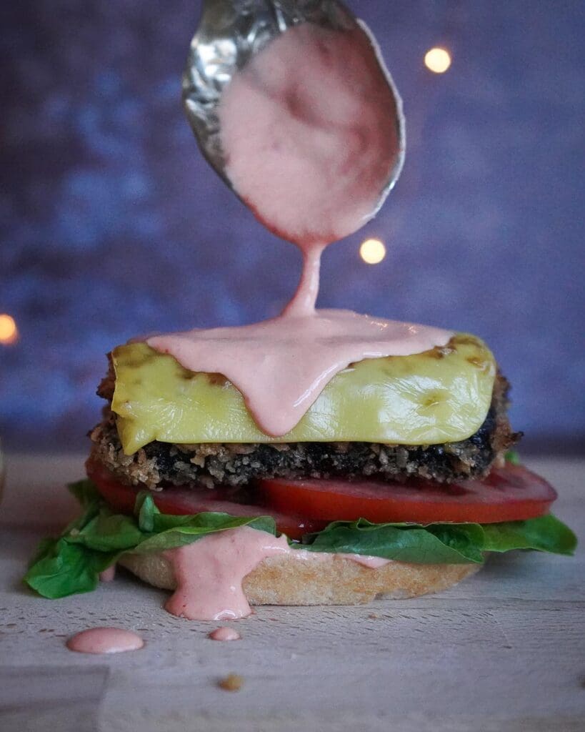 A fillet-no-fish vegan fish burger sat on a bed of lettuce and tomato, topped with melty vegan cheese and having chilli mayo drizzled over it