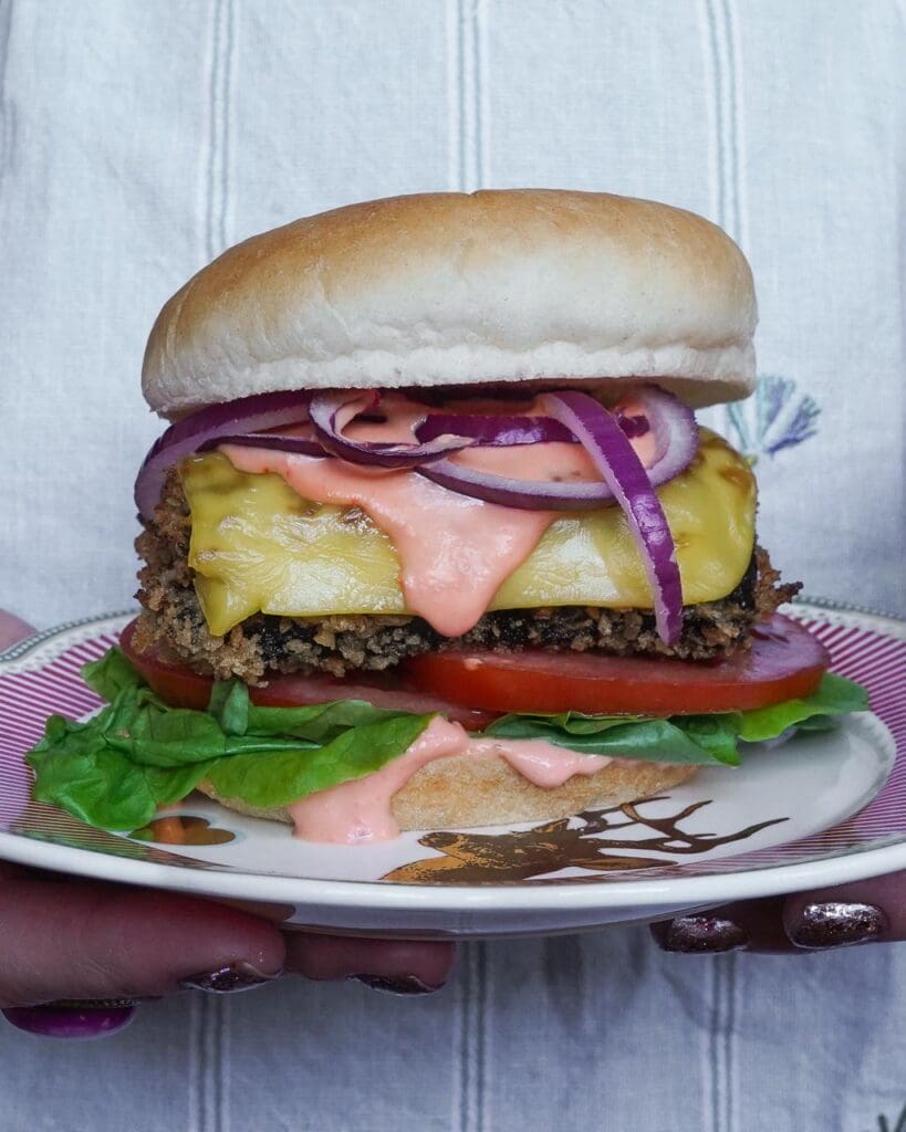 A stacked vegan fish burger with melty cheese, chilli mayo, red onion, lettuce and tomato in a soft bun