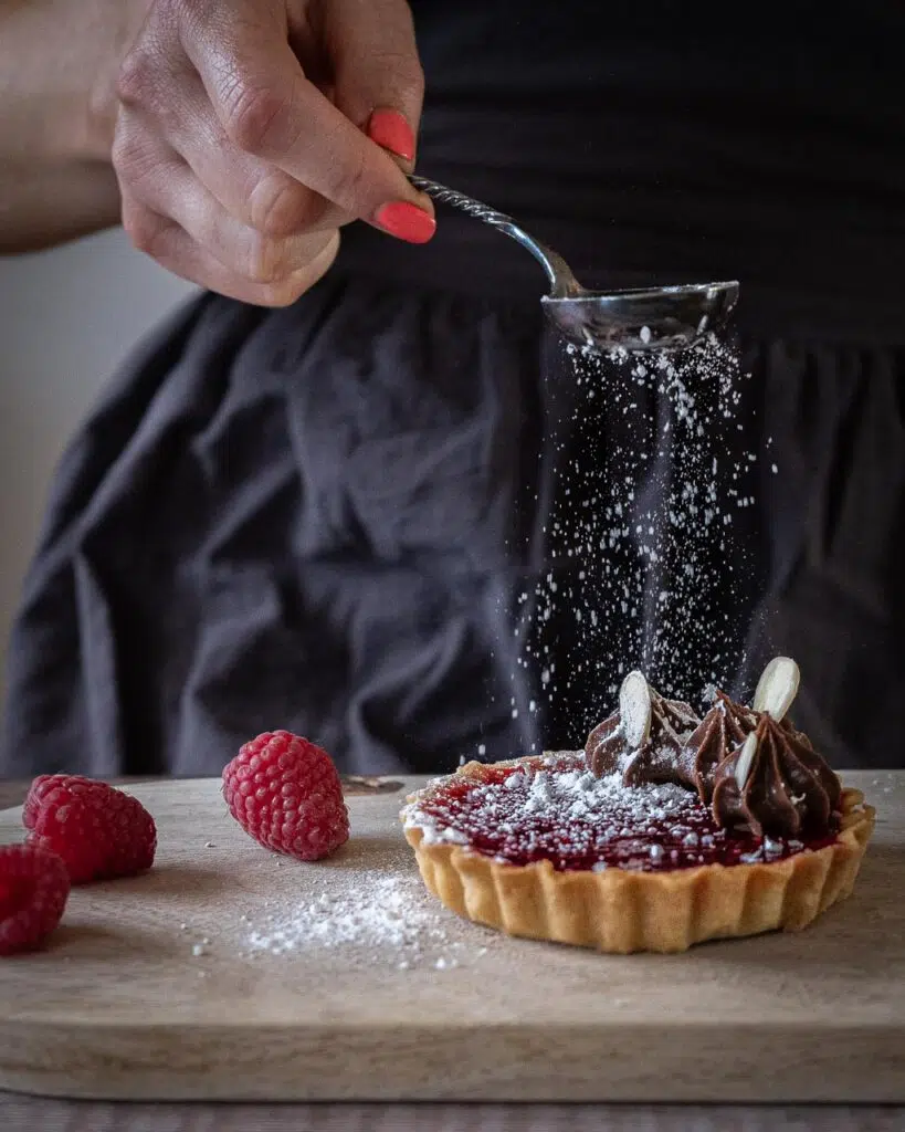 A raspberry vegan jam tart topped with piped chocolate cream and being dusted with icing sugar