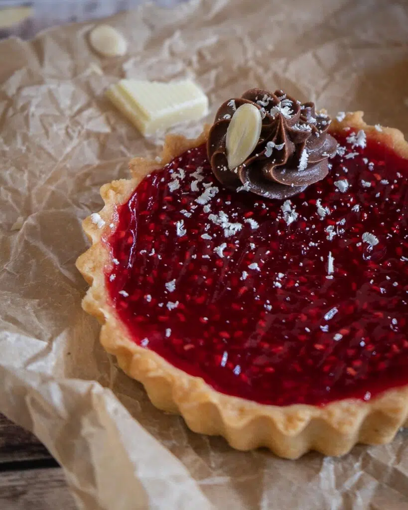 Close up of a vegan raspberry jam tart in a golden pastry case with piped chocolate cream on top and a sprinkling of grated white chocolate