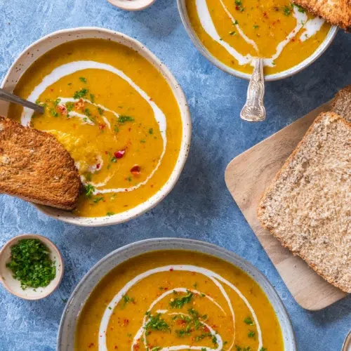 An overhead photograph of three bowls of red lentil and apricot soup. With a side of toast, a swirl of coconut milk and a sprinkling of red chilli.