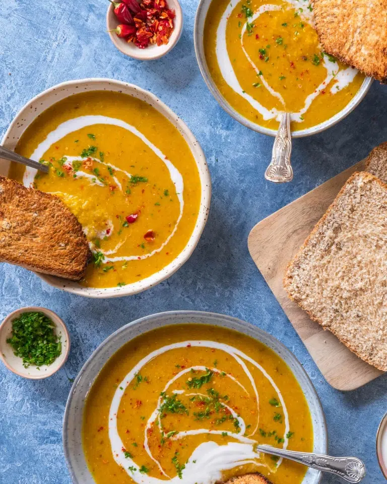 An overhead photograph of three bowls of red lentil and apricot soup. With a side of toast, a swirl of coconut milk and a sprinkling of red chilli.