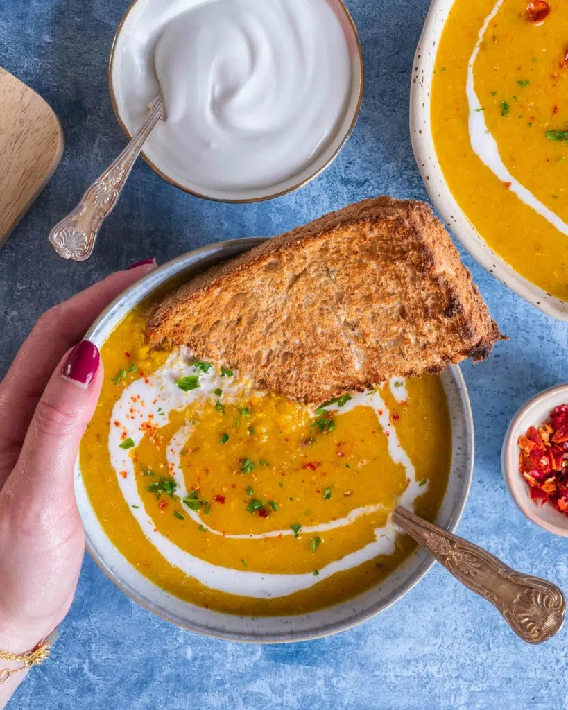 A warming bowl of Armenian Red Lentil Apricot Soup being held by a woman's hand.  The bowl has a slice of toast dipped in it and is swirled with coconut cream, red chilli and herbs.