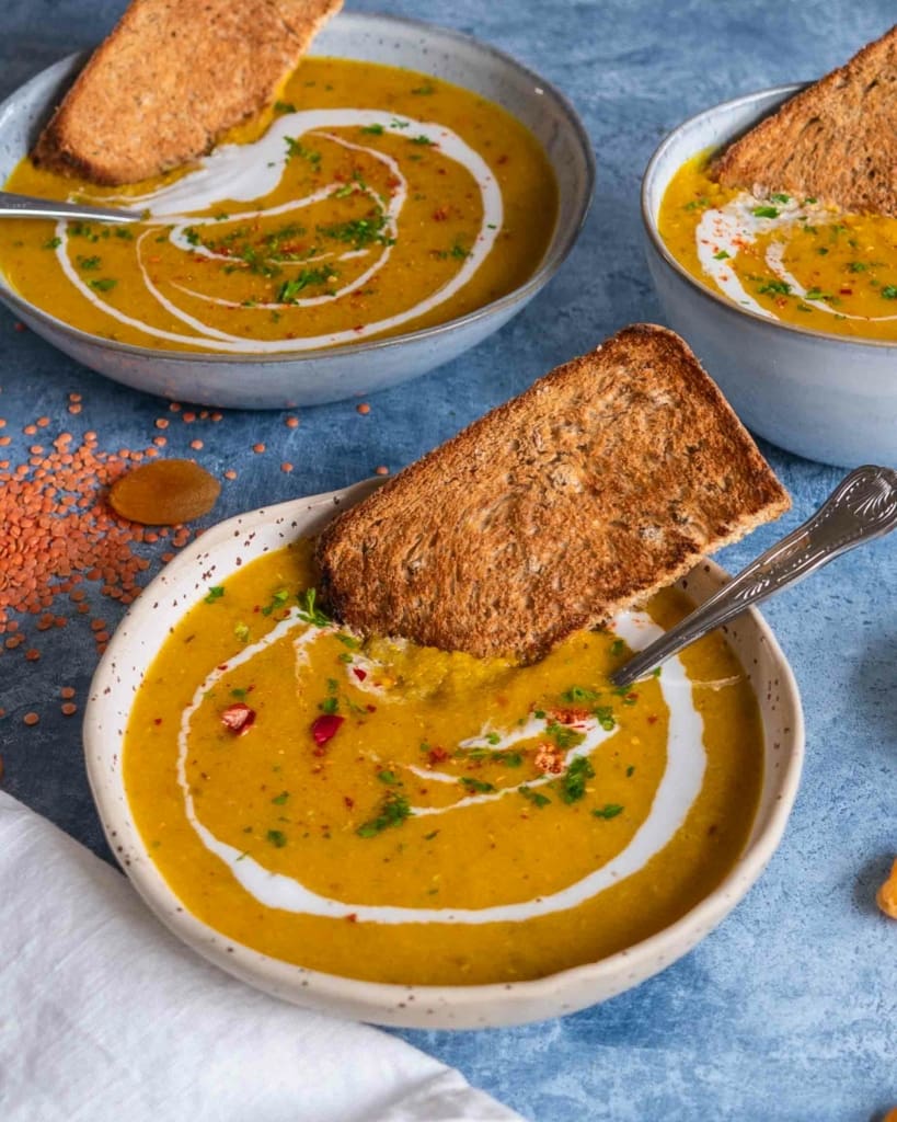 A side on photograph of three bowls of apricot lentil soup, swirled with coconut cream and sprinkled with red chilli.