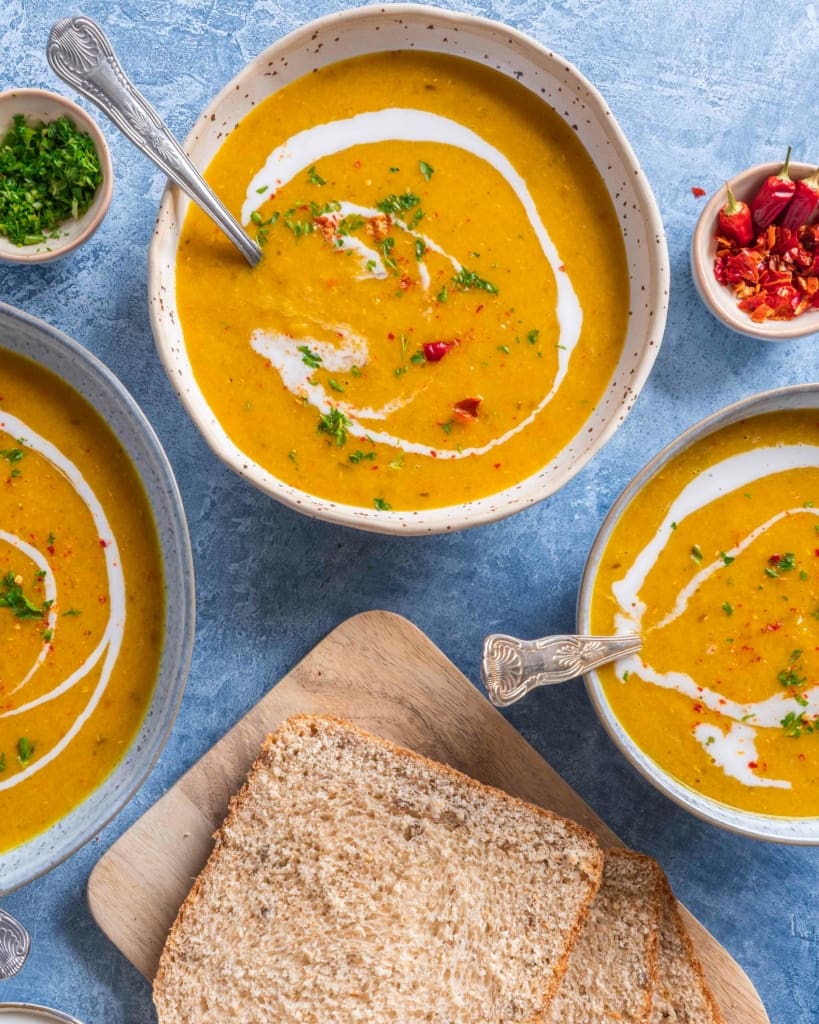 An overhead photograph of three bowls of Armenian Red Lentil Apricot Soup. With a side of toast, a swirl of coconut milk and a sprinkling of red chilli.