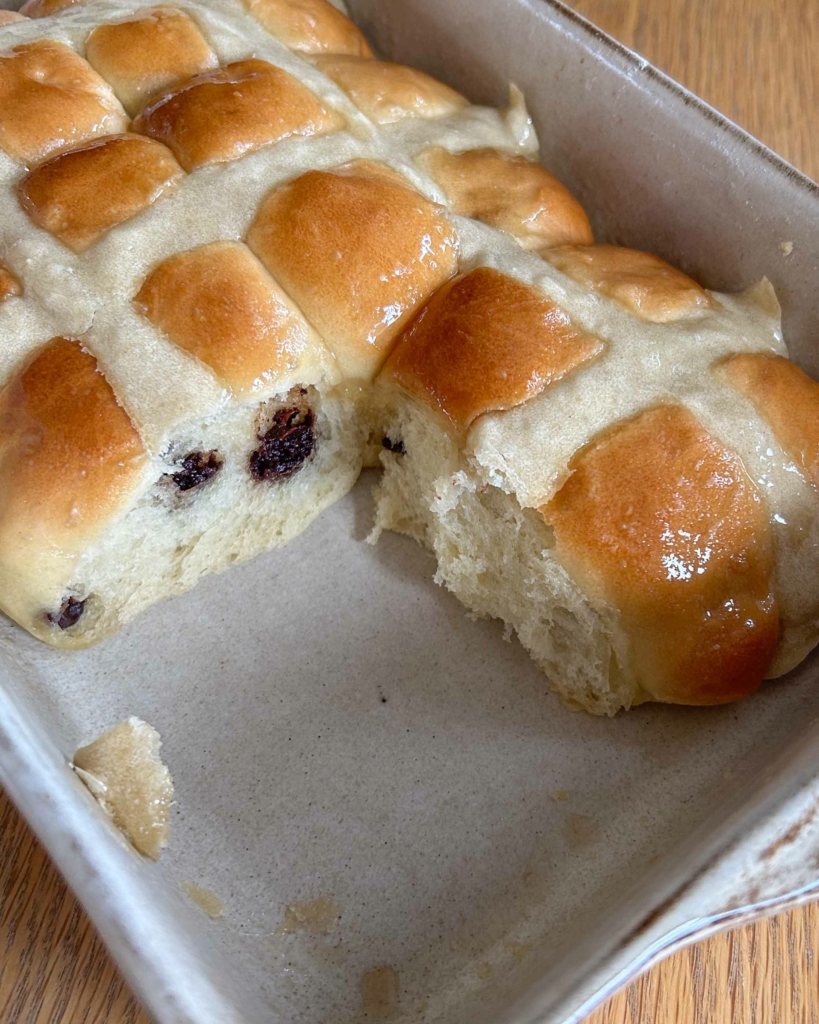 A dish of freshly baked vegan chocolate chip hot cross buns, with one bun torn out, revealing the soft and fluffy interior of the buns beside it