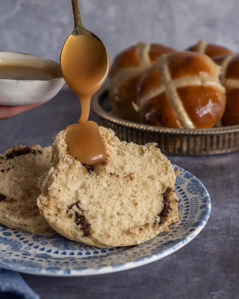 A soft and squishy vegan hot cross bun cut in half on a pretty blue plate, being drizzled with a salted caramel sauce