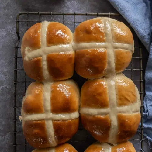 Soft and squishy vegan chocolate chip hot cross buns sat on a cooling rack with a glazed, golden top.
