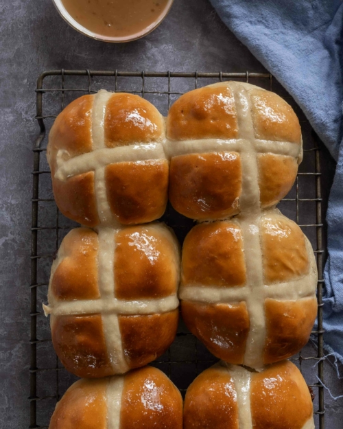 Soft and squishy vegan chocolate chip hot cross buns sat on a cooling rack with a glazed, golden top.