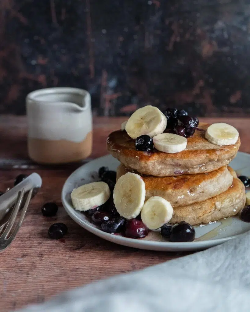 A stack of fluffy vegan pancakes with sliced banana, fresh blueberries and maple syrup