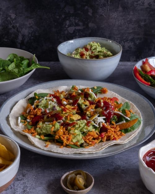 A vegan burrito in the making, with spicy rice, fajita vegetables, guacamole, chipotle sauce and crumbled tortilla chips, surround by small bowls of ingredients.