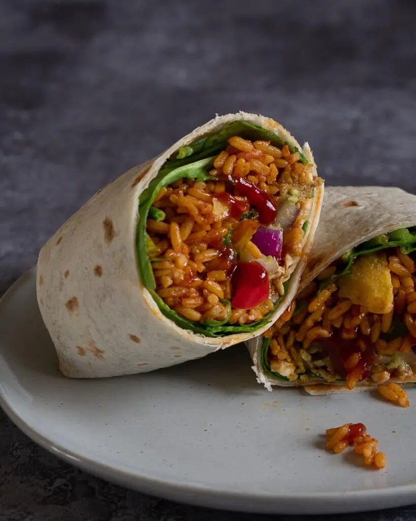 A vegan burrito cut in half with spicy rice, fajita vegetables, guacamole, chipotle sauce and crumbled tortilla chips wrapped in a soft tortilla wrap