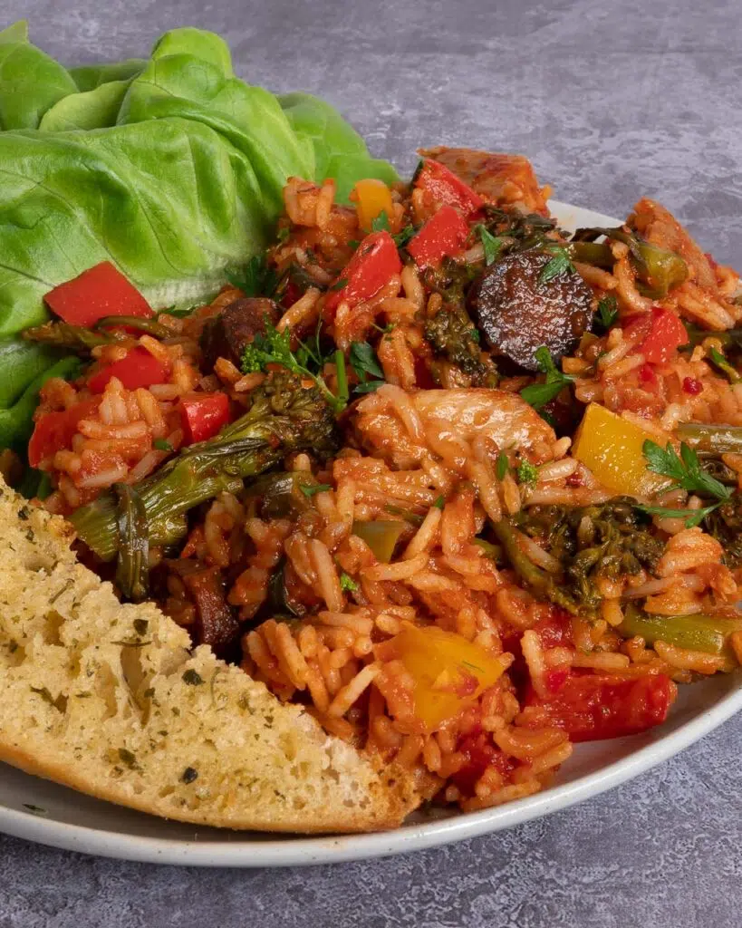 A bowl of colourful vegan jambalaya with orange rice, yellow, red and green vegetables, vegan chorizo and chicken and a side of garlic bread