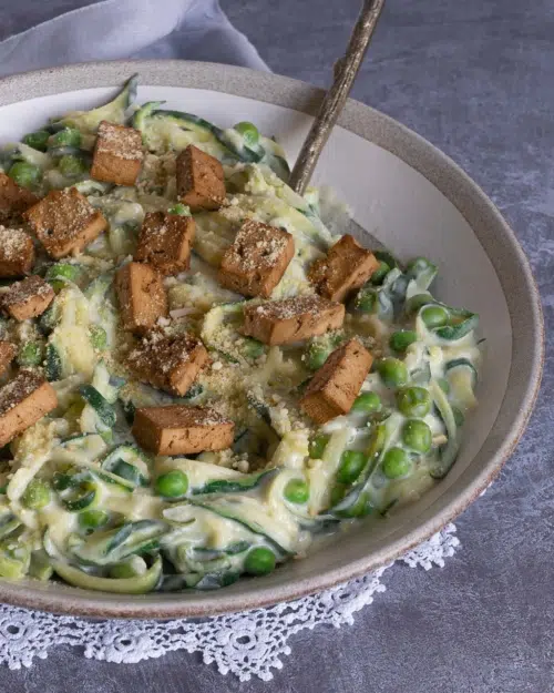 A bowl of healthy vegan carbonara made with zoodles in a creamy sauce, topped with smokey tofu bacon and vegan parmesan