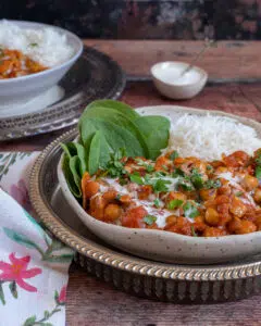A rustic bowl filled with a deep orange chana masala curry with a side of spinach and white rice and a garnish of fresh herbs and cream
