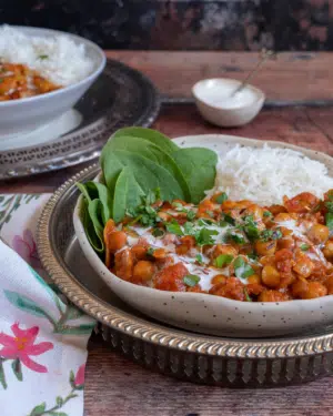 A rustic bowl filled with creamy chana masala curry with a side of spinach and white rice and a garnish of fresh herbs and cream