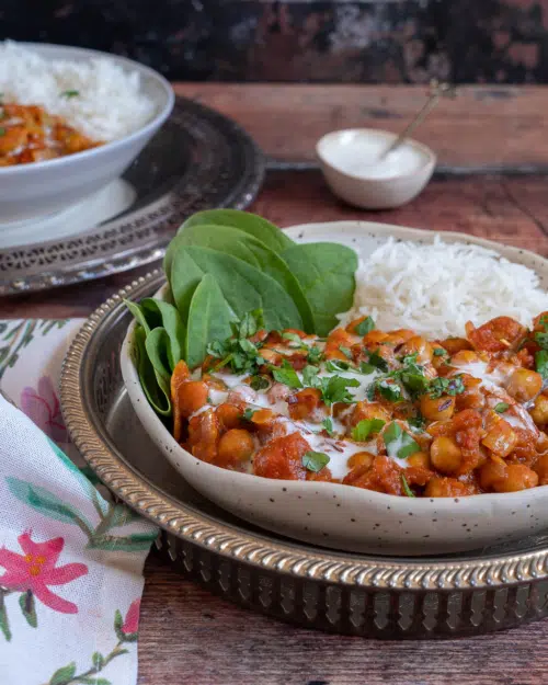 A rustic bowl filled with creamy chana masala curry with a side of spinach and white rice and a garnish of fresh herbs and cream