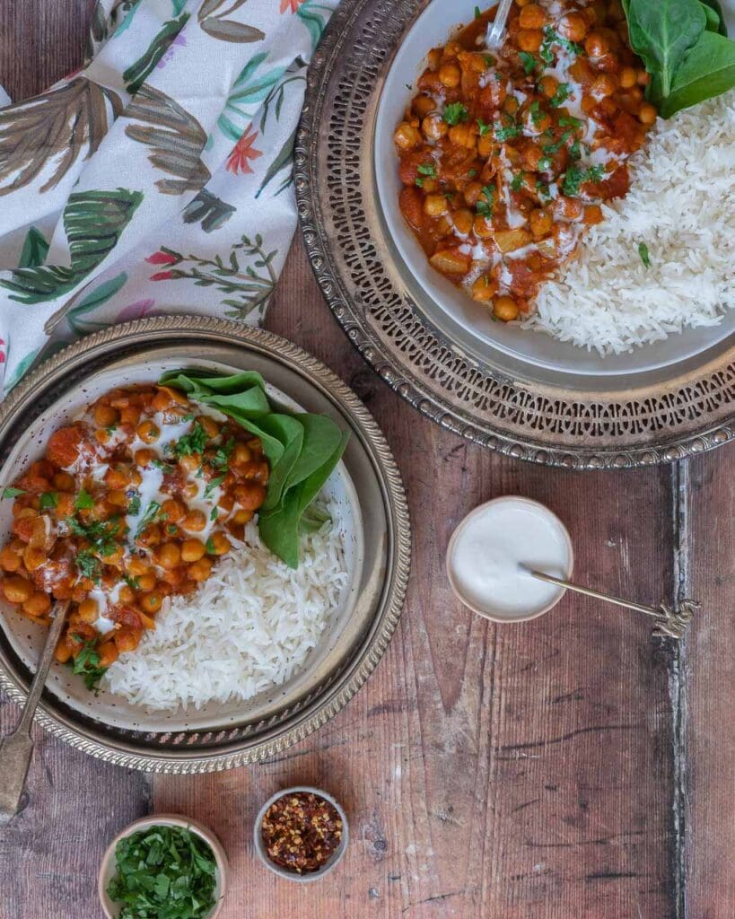 Top down shot of two bowls filled with a creamy chana masala curry with a side of spinach and white rice and a garnish of fresh herbs and cream