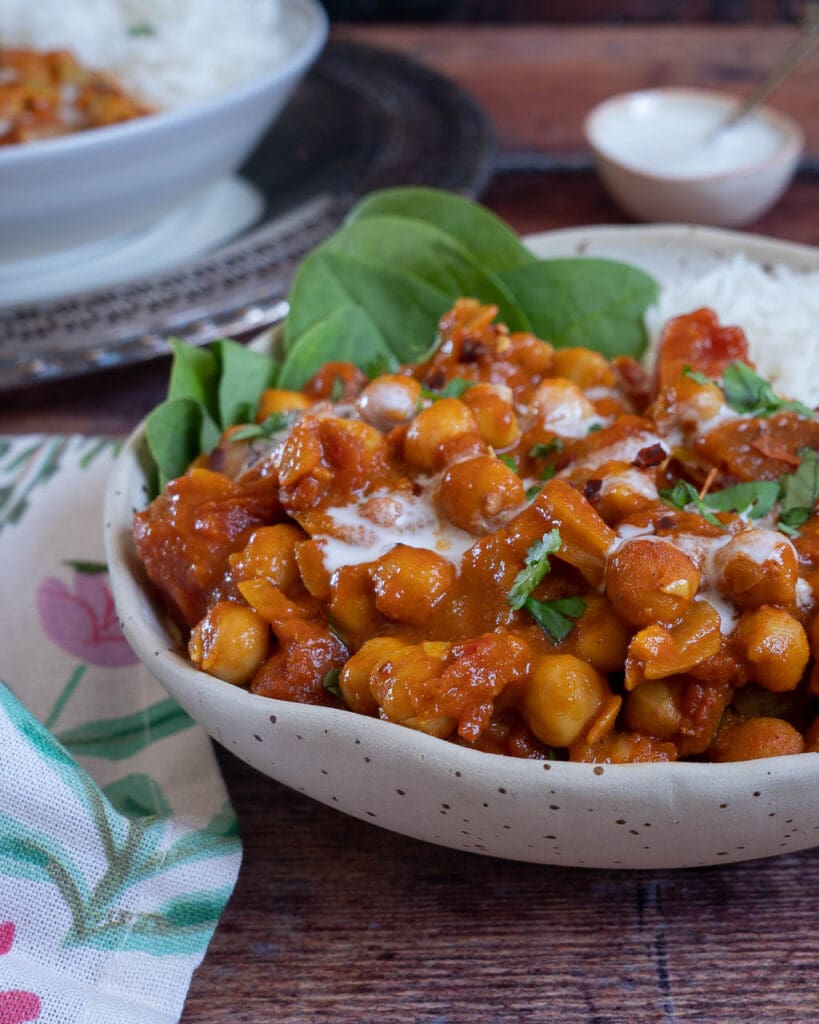 A rustic bowl filled with a deep orange chana masala curry with a side of spinach and white rice and a garnish of fresh herbs and cream