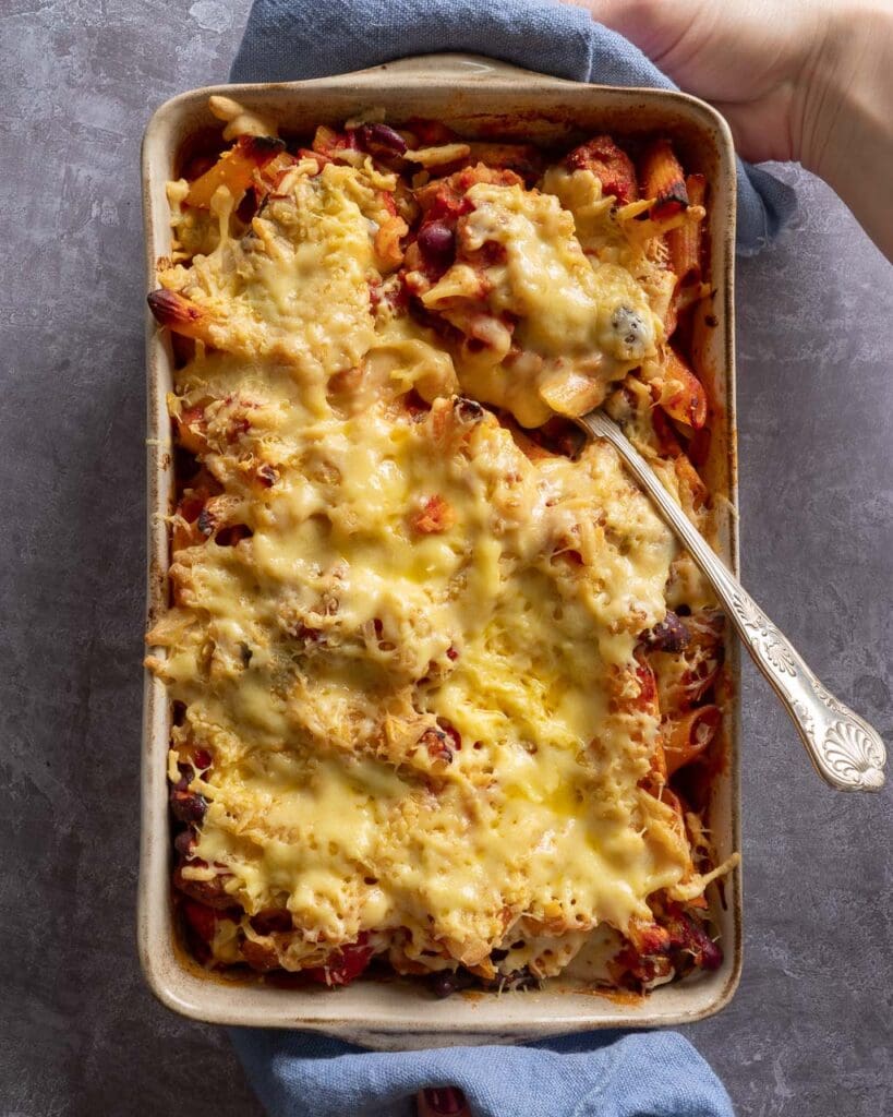 A baking dish filled with cheesy vegan sausage and bean pasta bake, with a serving spoon poised to scoop out a portion