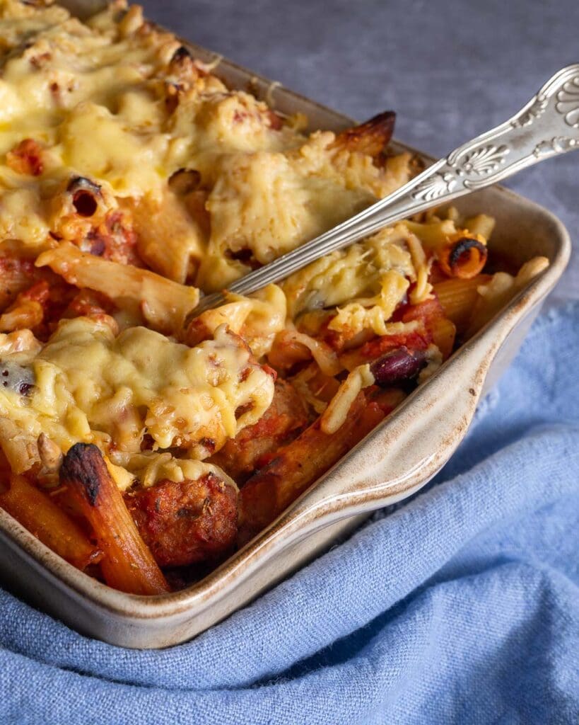 A baking dish filled with cheesy vegan sausage and bean pasta bake, with a serving spoon poised to scoop out a portion