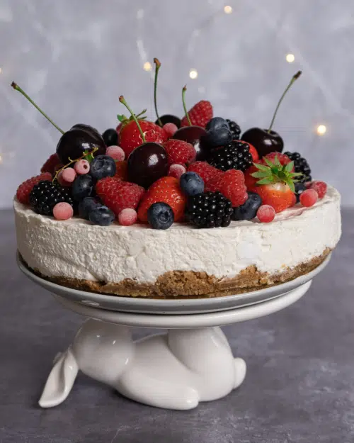 A no bake vegan cheesecake on a white rabbit stand, garnished with a pile of fresh berries