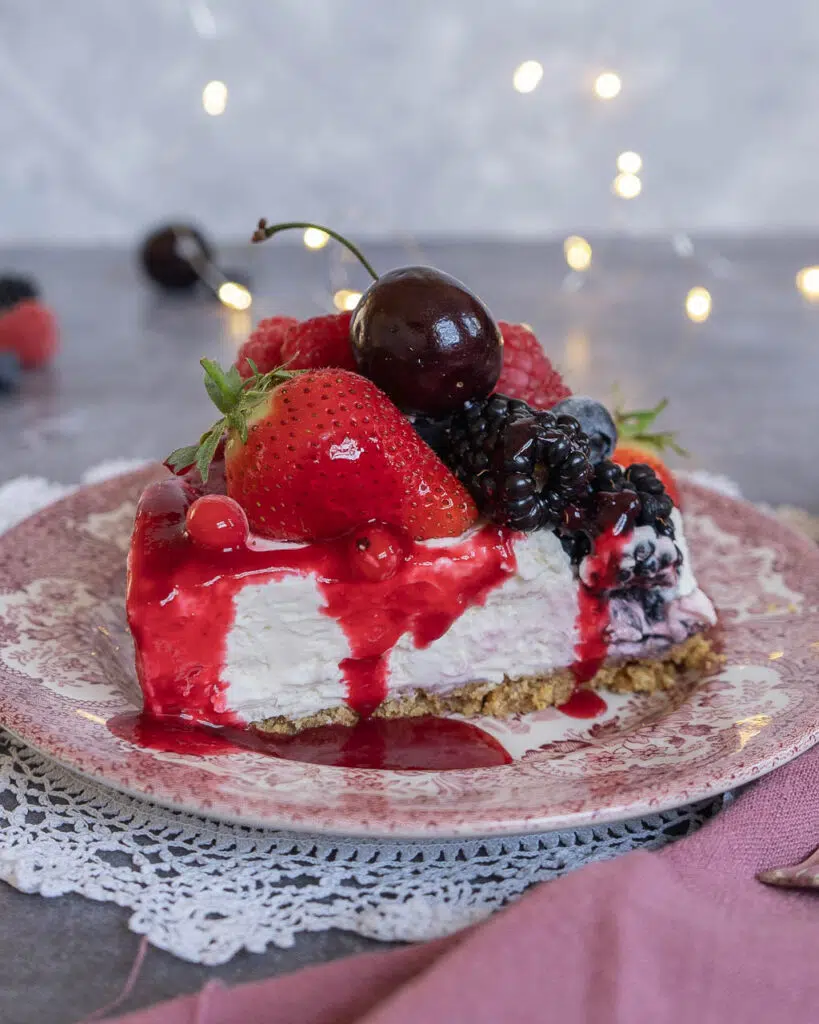A slice of creamy no bake vegan cheesecake on a pink and white patterned plate, topped with fresh berries and berry coulis