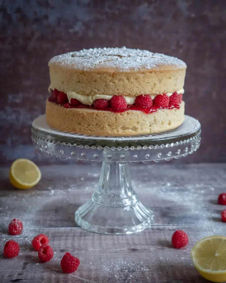 A vegan lemon cake on a glass stand, sandwiched with lemon buttercream and fresh raspberries