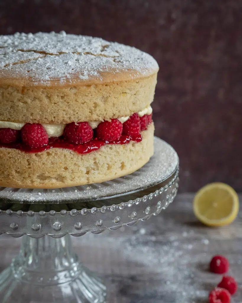 A vegan lemon cake on a glass stand, sandwiched with lemon buttercream and fresh raspberries