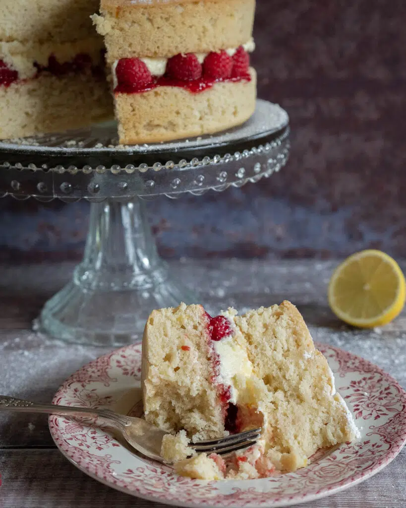 A slice of vegan lemon cake sandwiched with lemon buttercream, fresh raspberries and raspberry jam, sat on a pretty pink and white patterned plate