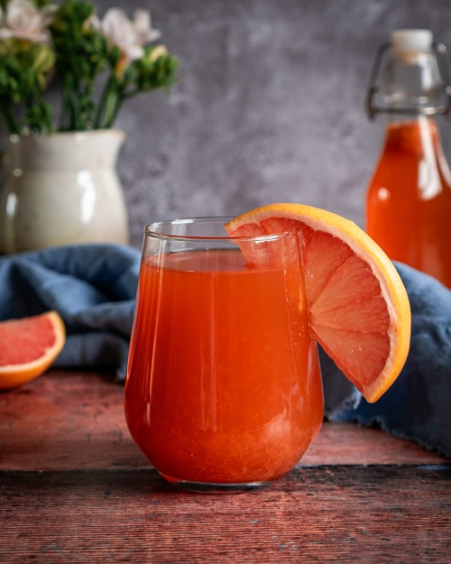 A fresh and bright glass of pink grapefruit kombucha sat on a tabletop with a slice of fresh grapefruit on the side of the glass