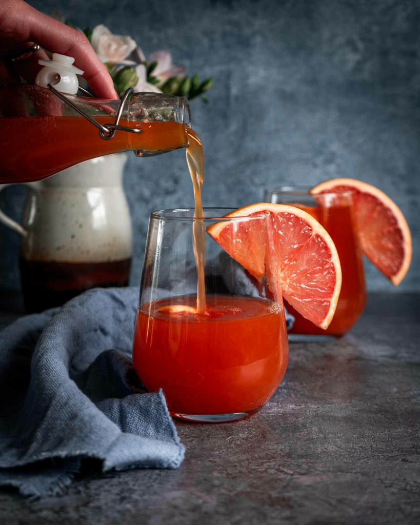 A glass of pink grapefruit kombucha being poured from a clip top bottle, with a slice of fresh grapefruit on the side of the glass