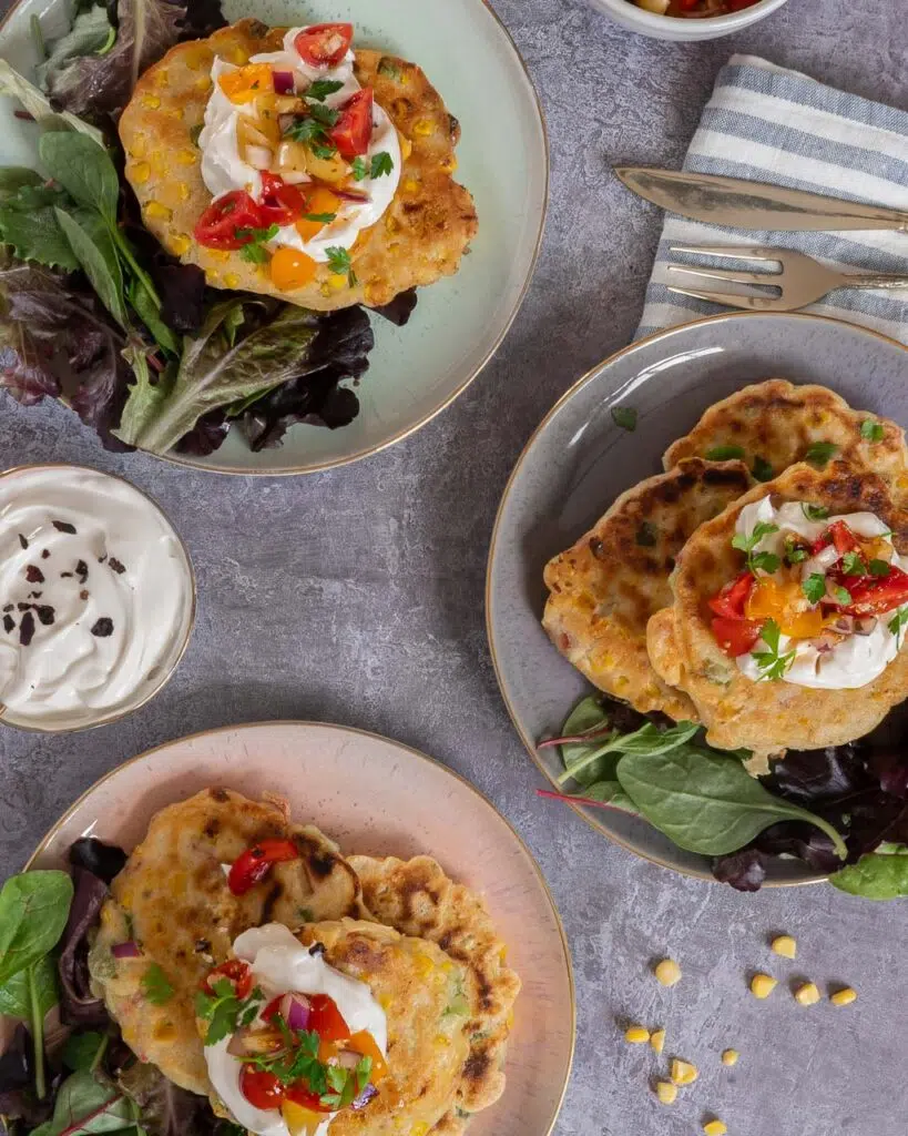 Top down view of three colourful plates of vegan sweetcorn fritters topped with creme fraiche and chipotle salsa