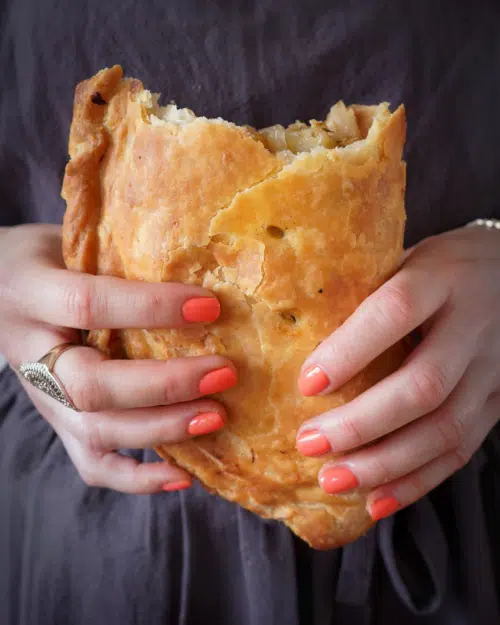 Two hands holding a massive vegan Cornish pasty with flaky pastry and the top broken off exposing the delicious filling