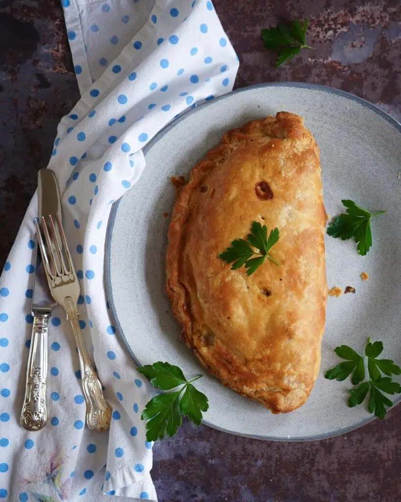 A large vegan Cornish pasty on a plate, with flaky pastry and garnished with fresh parsley leaves