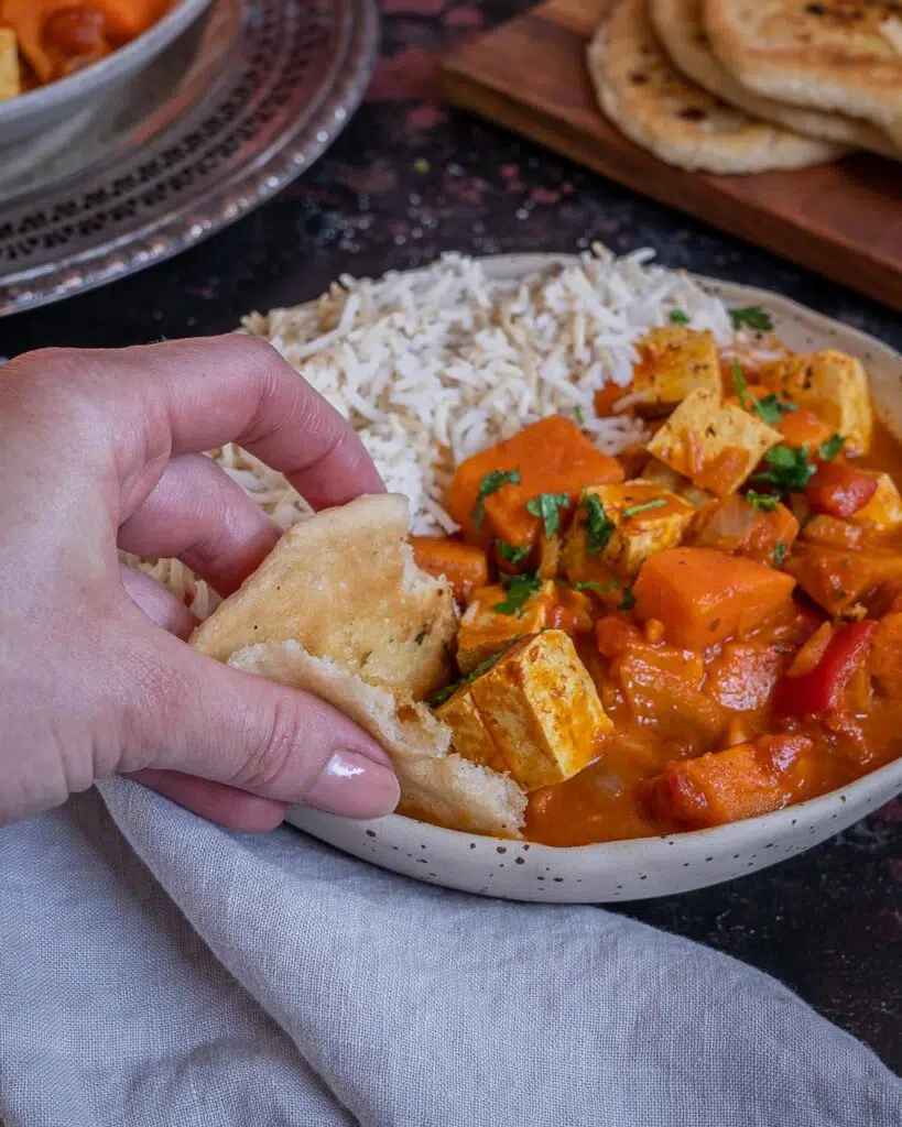 A bowl of rich orange vegan tikka masala served with white rice and naan bread being dipped into the sauce