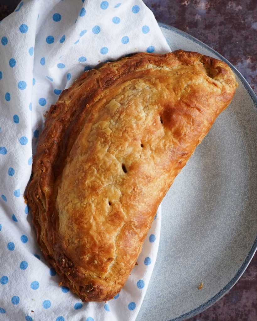 A beautifully golden, flaky vegan Cornish pasty on a plate with a dotty napkin beside it