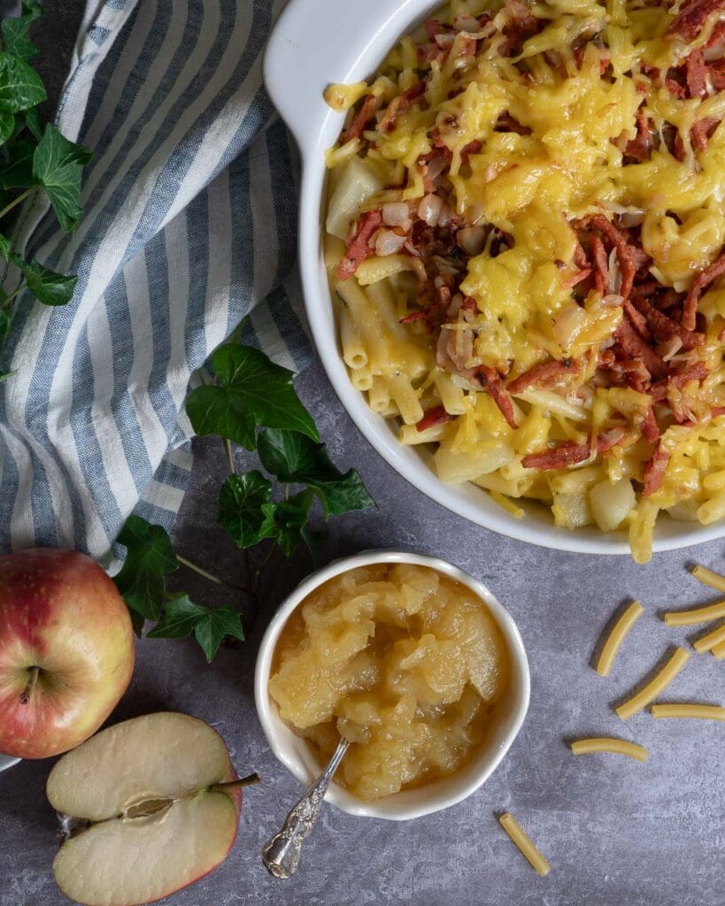 A large dish of vegan mac n cheese topped with veggie bacon, melted cheese and served with a side of apple sauce