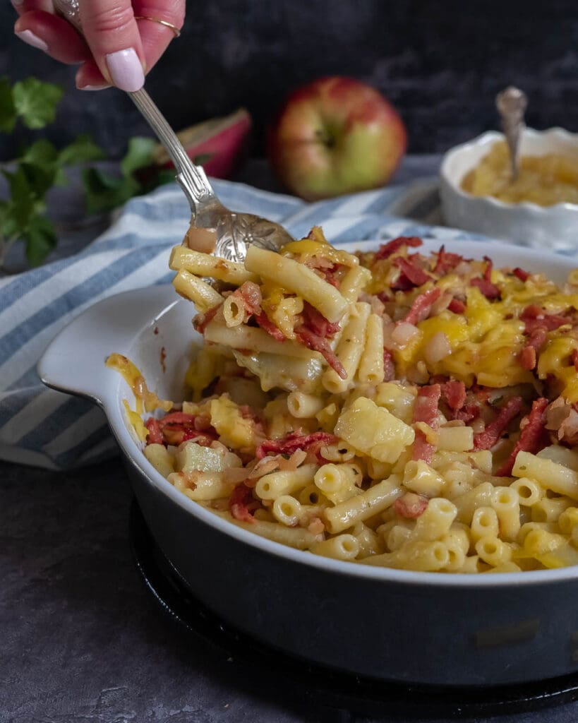 A large dish of vegan mac n cheese topped with veggie bacon, melted cheese and served with a side of apple sauce