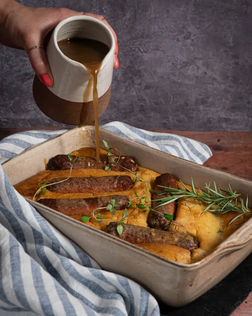 An oven dish filled with crisp and fluffy vegan toad in the hole, golden veggie sausages and fresh herbs being drizzled with vegan gravy