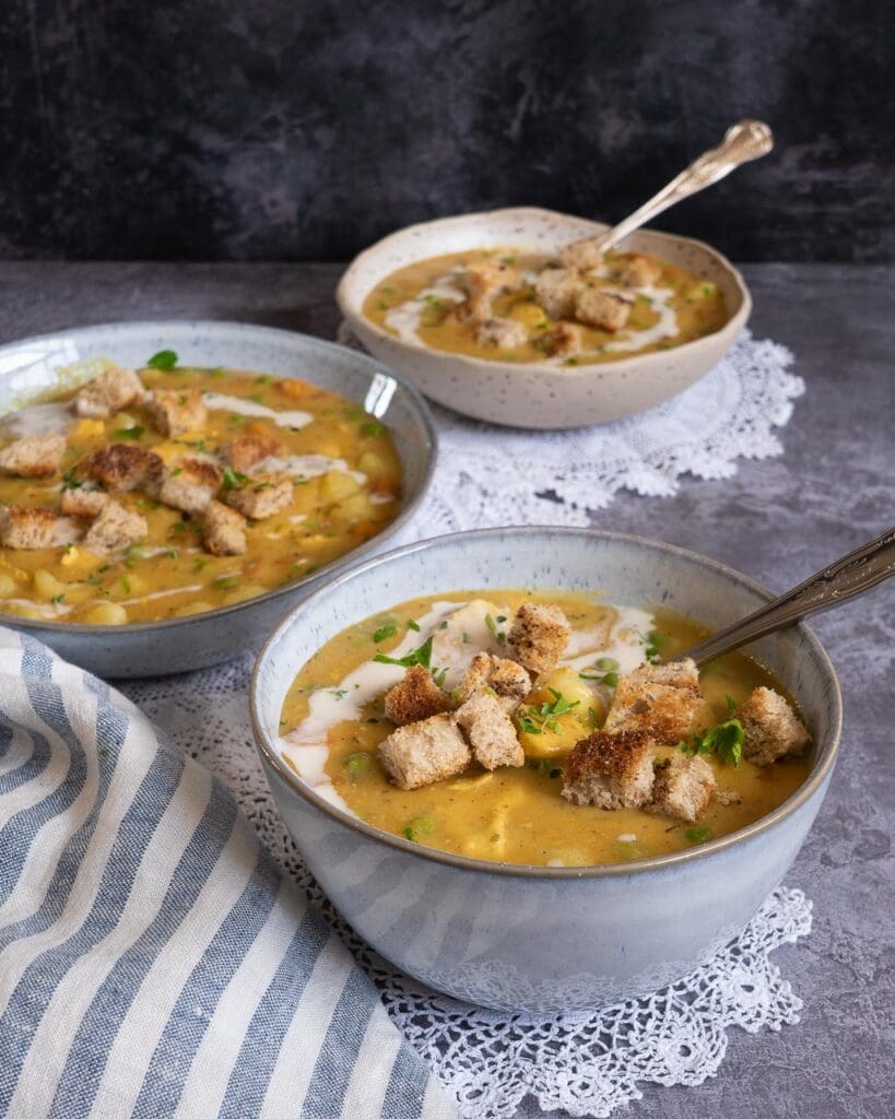 A side-on photograph of three bowls of vegan chicken pot pie soup. The table top is adorned with lace doilies and a striped tea towel. The soup is a rich golden colour and is topped with crunchy garlic croutons.
