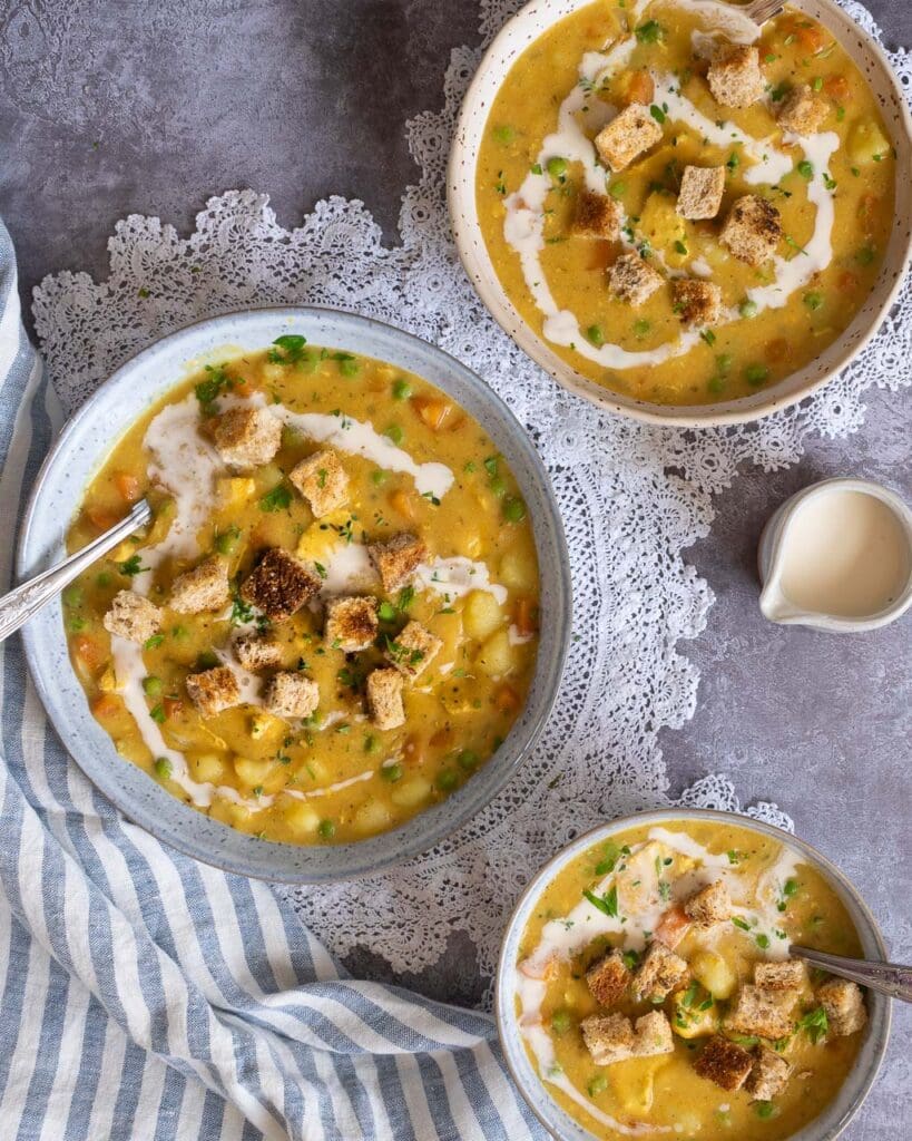 An overhead photograph of three bowls of vegan chicken pot pie soup. The table top is adorned with lace doilies and a striped tea towel. The soup is a rich golden colour and is topped with crunchy garlic croutons.