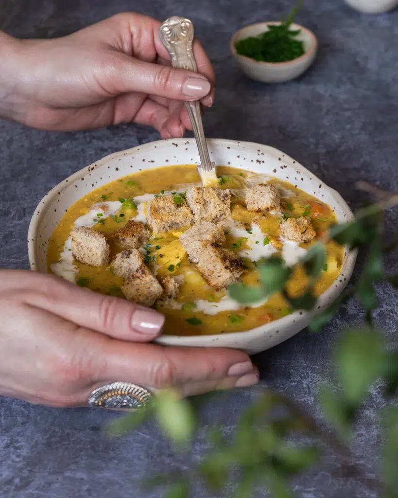 An up-close photograph of a woman stirring a rustic bowl of vegan chicken soup. There is a plant blurred out in the foreground, with the soup in focus; it's a golden colour with a swirl of white from the vegan cream, fresh vegetables and herbs visible through the surface of the soup.